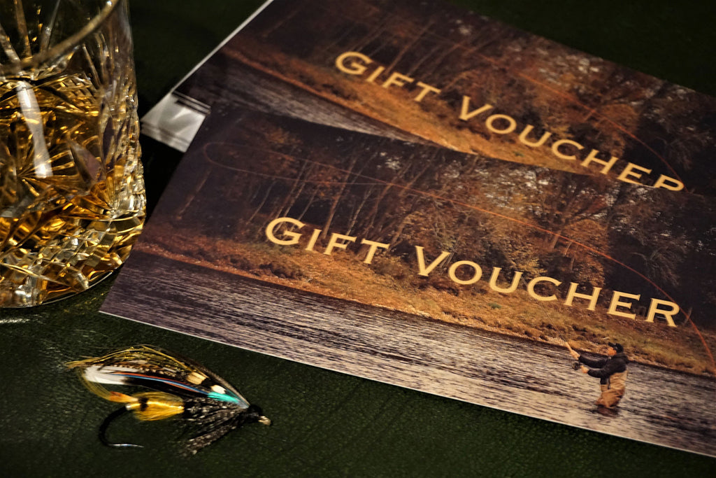 A gift voucher titled 'Gift Voucher' placed on a table with a glass of whiskey and a detailed fly fishing lure next to it, by Andrew Toft. 