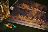 A gift voucher titled 'Gift Voucher' placed on a table with a glass of whiskey and a detailed fly fishing lure next to it, by Andrew Toft. 