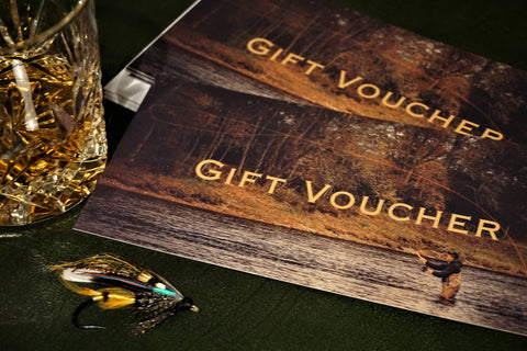 A gift voucher titled 'Gift Voucher' placed on a table with a glass of whiskey and a detailed fly fishing lure next to it, by Andrew Toft. 